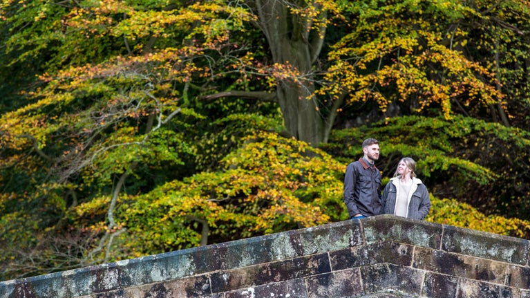 Man and woman standing side by side on arched stone bridge with autumn foliage in background